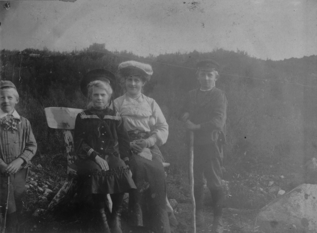 Ved Tårnelven 14. august 1907. En dame og en pike sitter på en benk. To gutter står på hver side av benken. Den ene holder en stokk i hånden.
