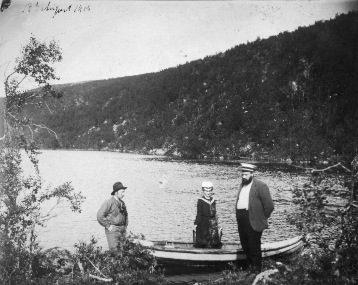 18. august 1906. En robåt ligger i vannkanten. En dame står i båten med en fisk i hver hånd. To menn står ved siden av båten. Det er trær rundt vannet.