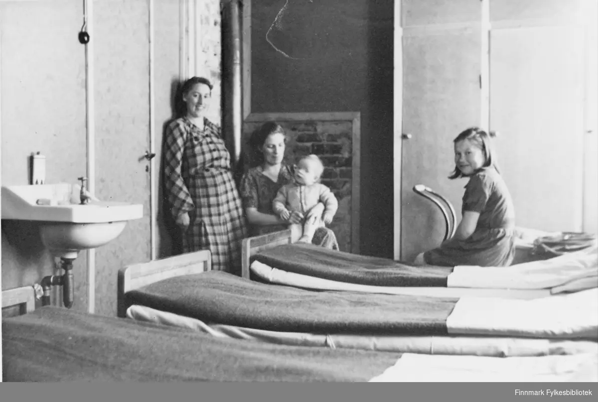 'Sykehus i Honningsvåg - en av sykestuene.' Et av rommene i sykestua. To kvinner og to barn, tre senger og en vask. Den permanente sykestua stod ferdig i 1950. Norges Røde Kors stod for driften til 1952/53 da Interesseselskapet overtok.