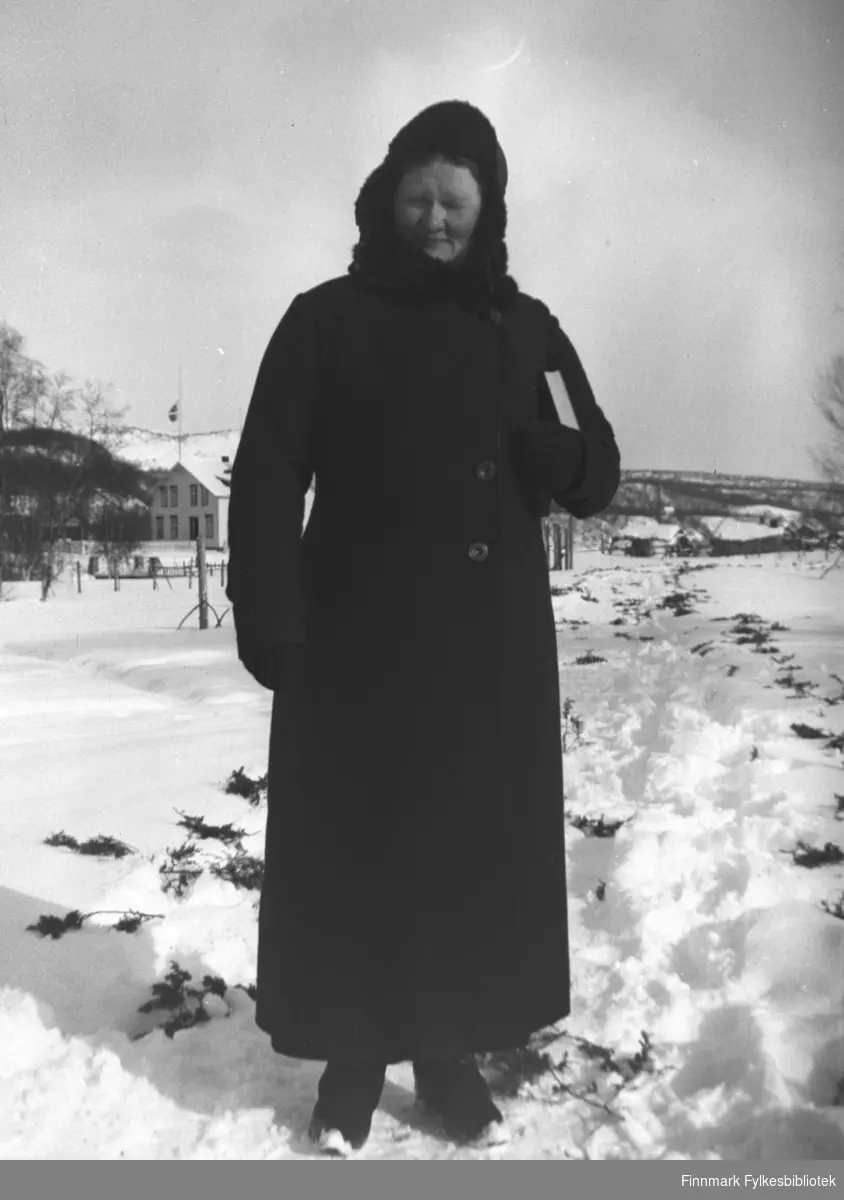 Kathinka Mikkola kledd i mørk vinterkåpe og skinnlue. Hun har salmeboka under armen. Bildet er tatt ved kapellet i Neiden i anledning en begravelse. Langs veien ligger einerkvister, kanskje i anledning begravelsen