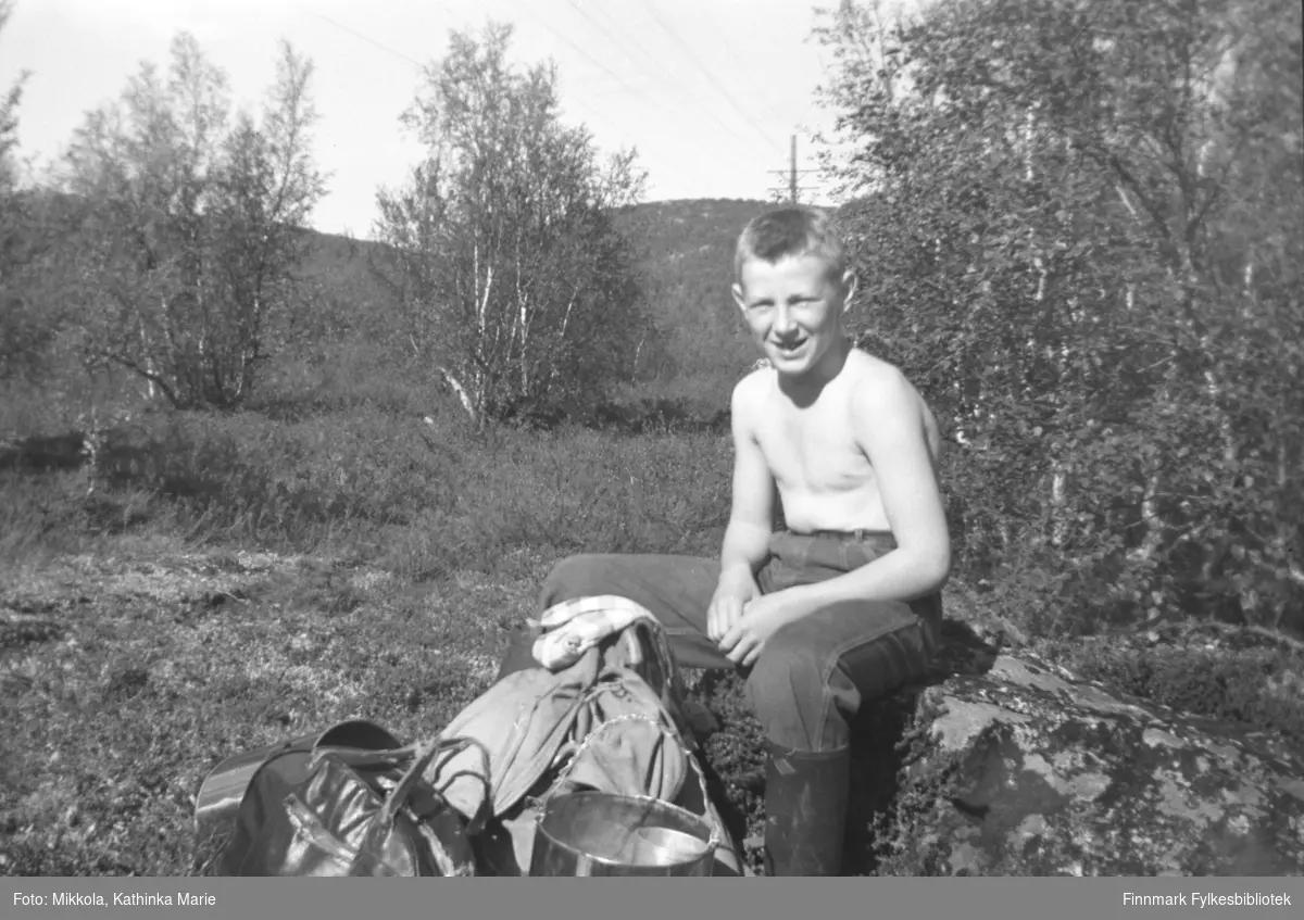 Willy Olsen på bærtur i Neiden-distriktet. Han sitter med bar overkropp på en stein og smiler mot fotografen, og foran ham ser vi ryggsekk og bærspann