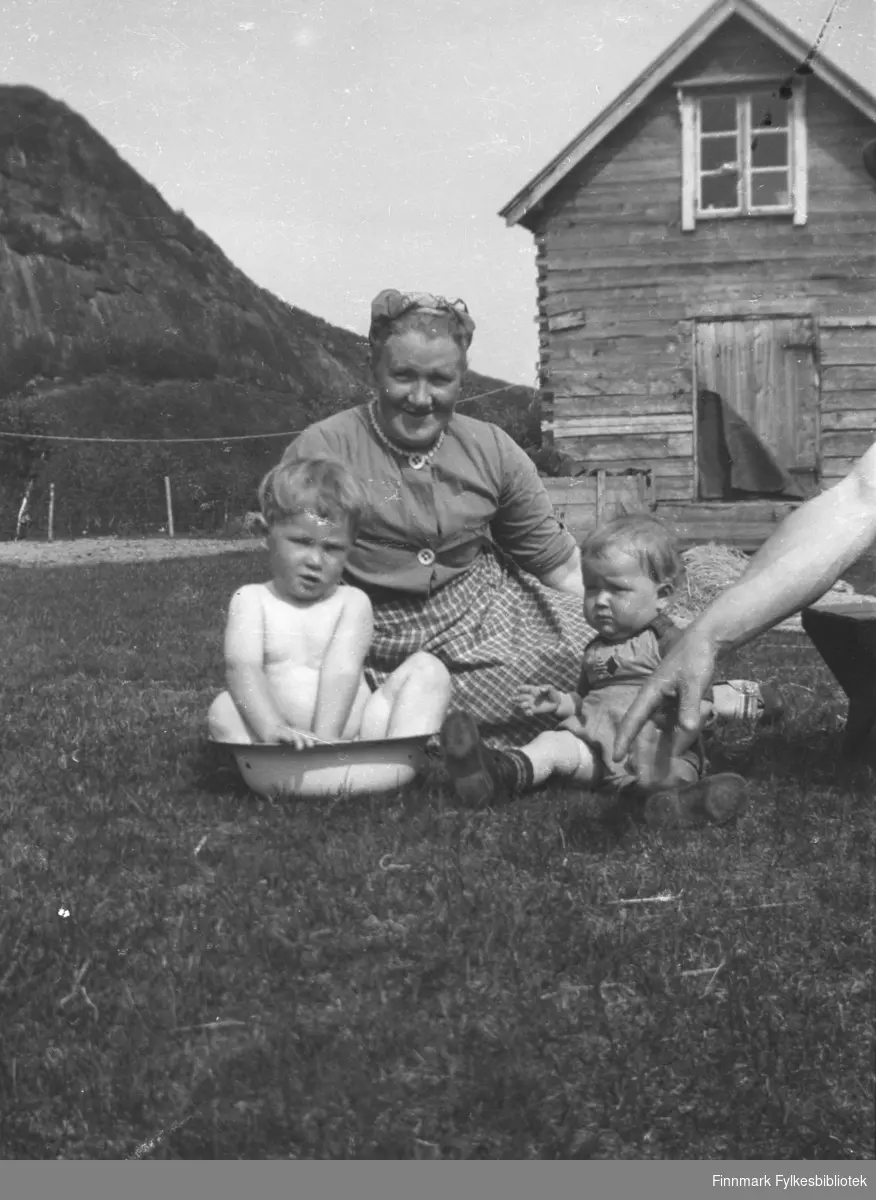 Willy Olsen bader i et vaskevannsfat på gårdsplassen på Mikkelsnes. Bak ham sitter hans mor, Synnøve Olsen, og den lille jenta heter Grete Lill Olsen. En naken mannsarm kommer uventet inn på motivet fra høyre kant. I bakgrunnen melkebua på gården.