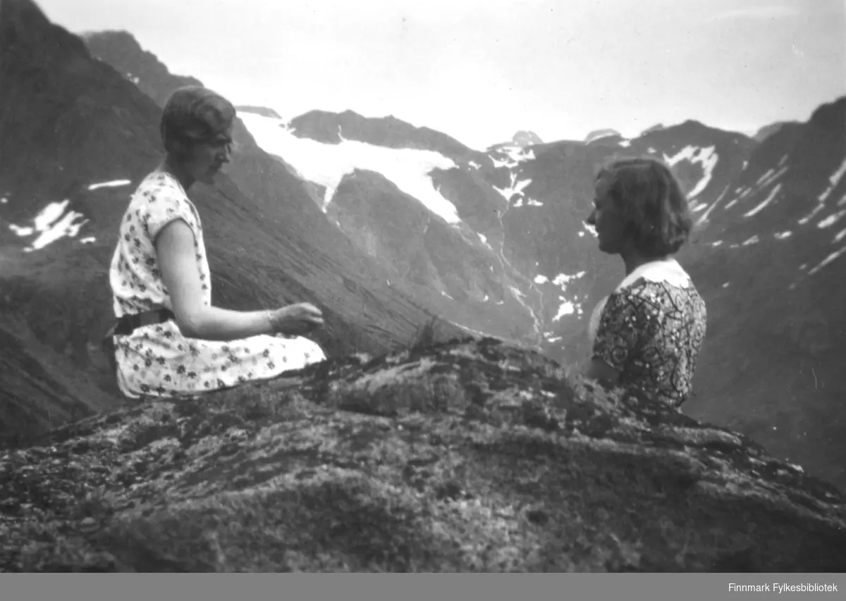 To damer sitter på en fjelltopp. Hun til venstre har en kortermet mønstret kjole med sort belte. Hun til høyre har en mørk kjole med mønster og hvit krave. Det er stein og litt lyng/gress på toppen rett bak dem. I bakgrunnen er flere høye fjell med is-/snøflekker på toppene. En større is-/snøflekk midt på bildet er muligens deler av Øksfjordjøkelen.