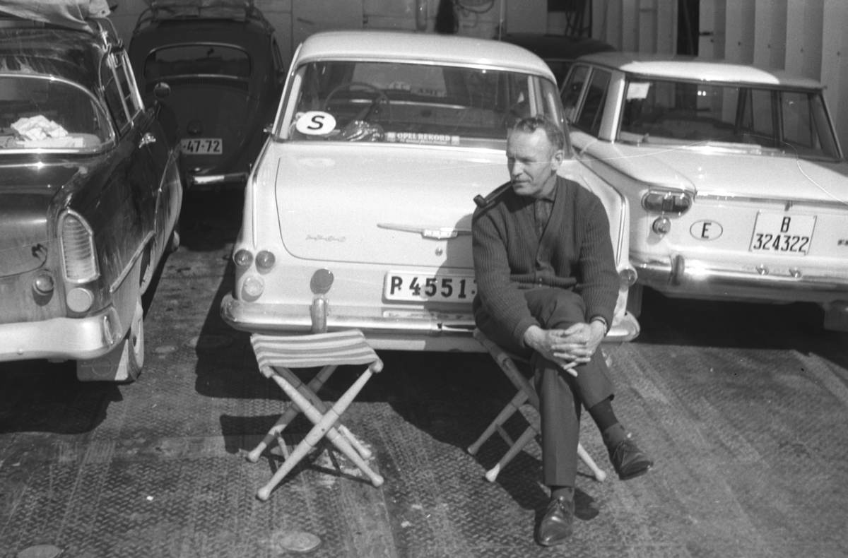 En ukjent mann sitter på en klappstol, muligens på et fergedekk. Bilen som står til venstre er en Opel Rekord P 1958-60 mod. Rett bak mannen står en lys Opel Rekord P2 1961-1962 mod. og til høyre står en Fiat 1300/1500, kan også være en Seat. I bakgrunnen kan hekken på en VW Boble ses.