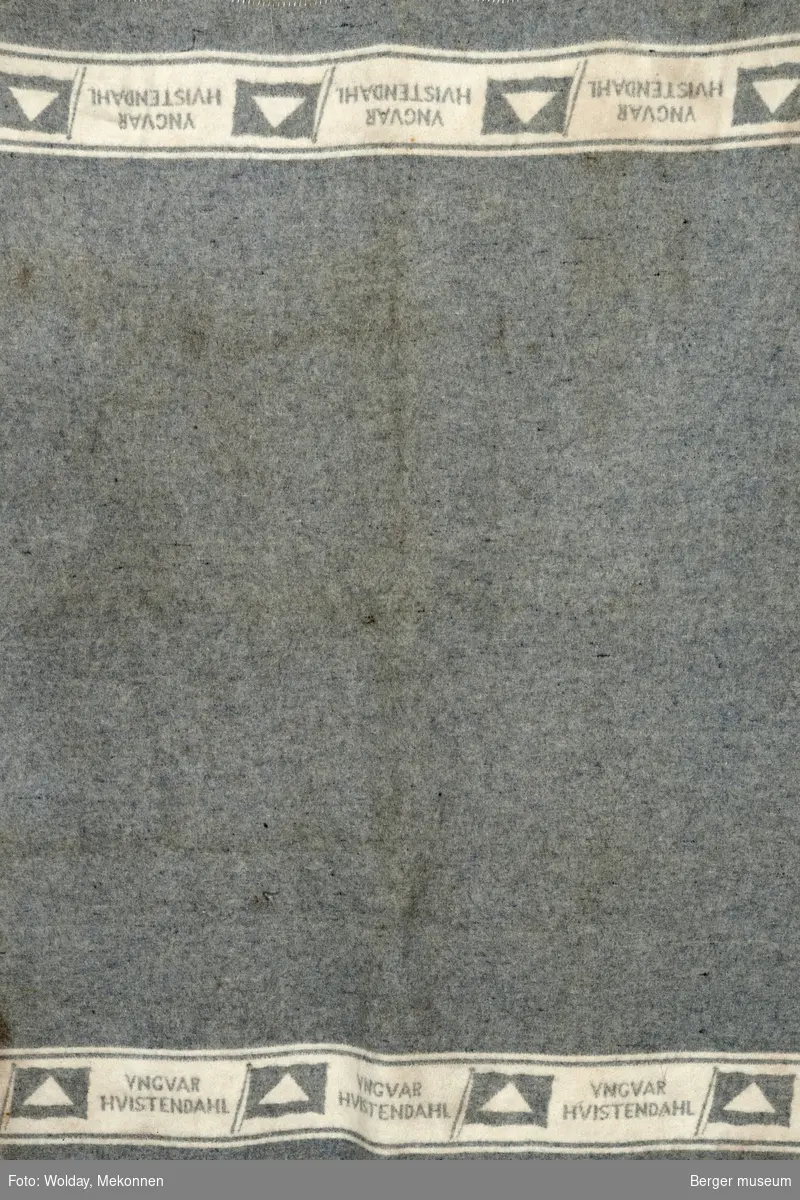 Grått teppe med logo for Yngvar Hvistendahls rederi på hvit stripe i hver ende av teppet. tekstfelt invertert på andre siden av teppe