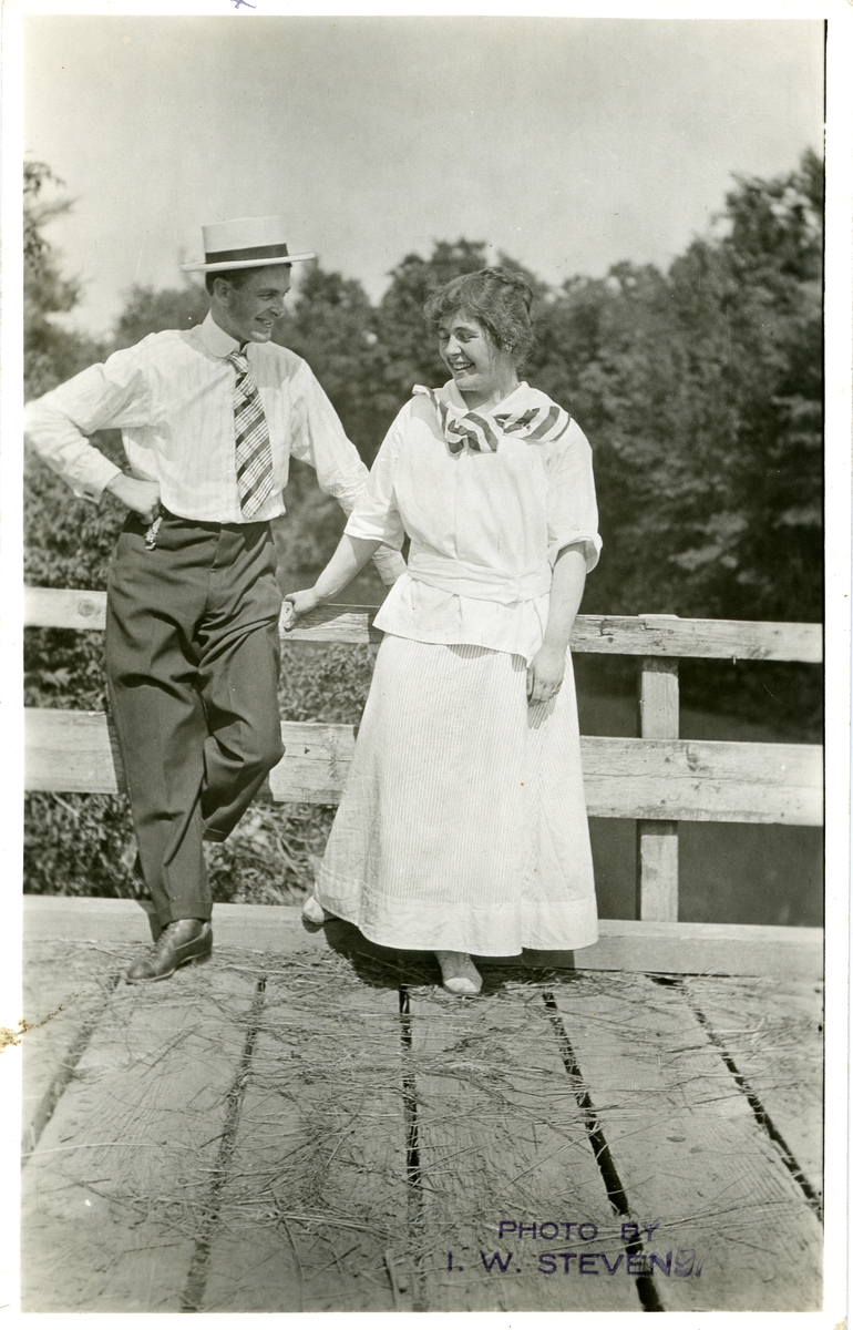 Mann og kvinne avbildet på ei trebru. Hun har langt hvitt skjørt og bluse. Han har mørtk buske, lys skjorte, slips og hatt