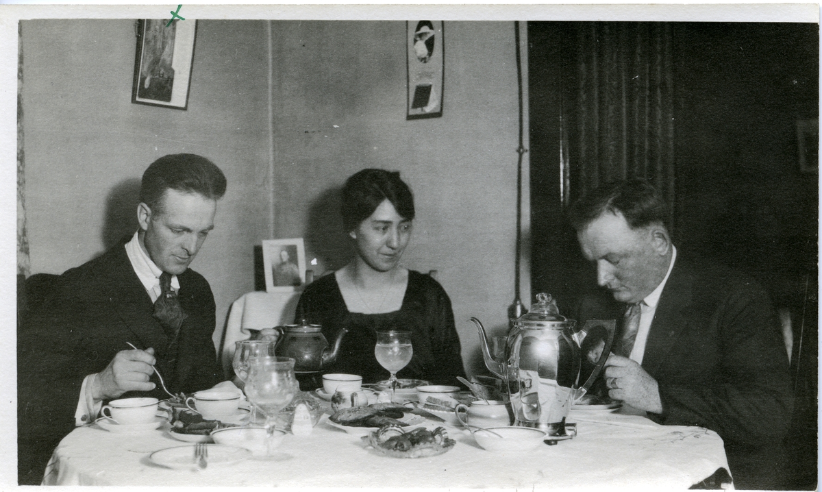 En kvinne og to menn sitter ved et pådekket spisebord. Kaffekopper, glass, kaker og kaffekanne på bodet.