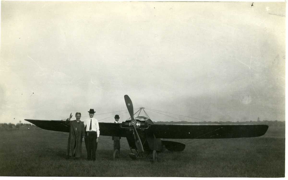To menn avbildet foran et fly. En tredje mann avbildet bak flyets vinge. To av mennene er iført dress og hatt mens den tredje er iført en form for kappe. Dette er et postkort.