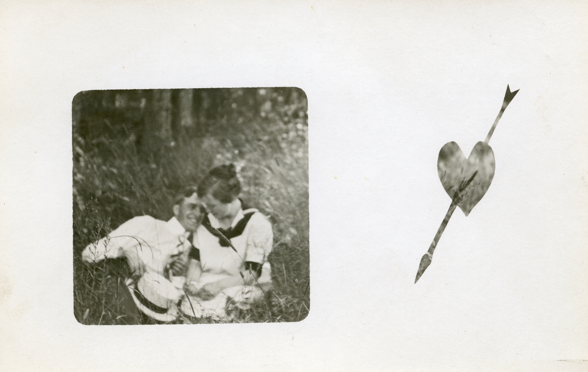 Postkort som viser to unge mennesker som sitter i gresset. Kortet har et hjertemotiv med en pil gjennom, til høyre for bildet.