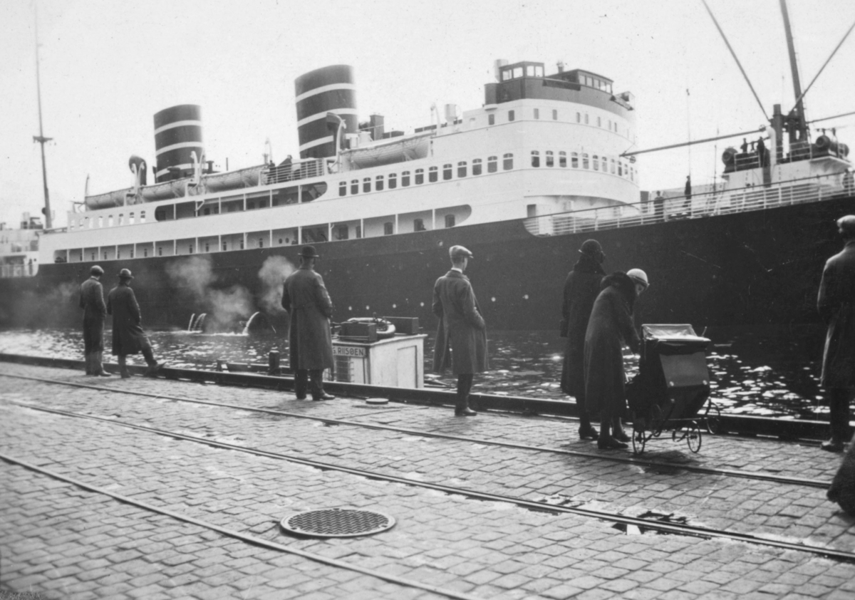 Skipet Venus ved kai i Bergen, 1932. Noen menn, og en kvinne med en barnevogn, står på den brosteinlagte kaien (veien)?