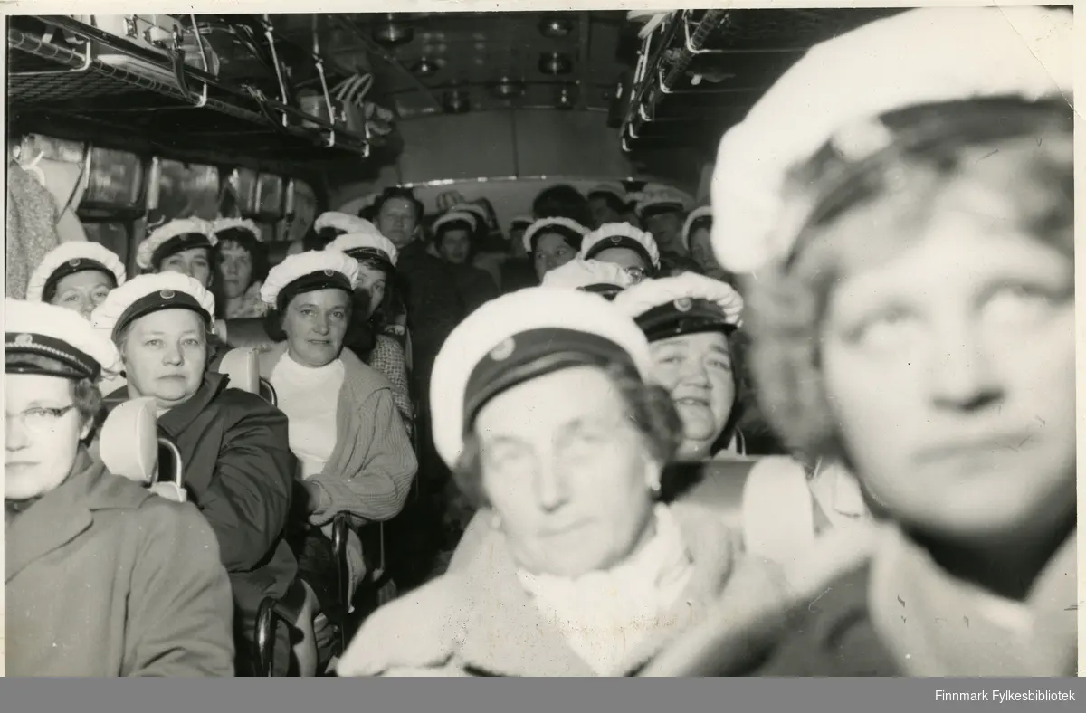 Vadsø damekor i bussen på tur til Berlevåg (muligens rundt 17. mai) 1962. Kvinnene har på seg en hvit hatt, trolig kor uniform. I bussens håndbagasje hylle kan man se vesker og kofferter av forskjellige slag. Damene har på seg jakker og gensere. Nederst i venstre hjørne: Ingrid Niskavara. Bak Bildet er det notert: 'M.Z', for Mimmi Zahl.