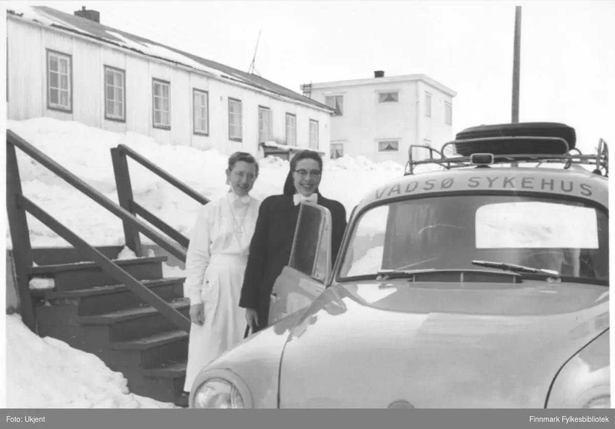 Reisende sykepleier Ingeborg Søvik står ved siden av en bil med tekst Vadsø sykehus (Opel Olympia varebil årsmodell 1953-57). Til venstre for henne står oversykepleier Gudrun Byholt. I bakgrunnen kan man se søsterbrakka. Gudrun har på seg uniform og et kors rundt halsen. Ingeborg har på seg jakke. De har begge briller. På taket på bilden kan man se bagasje. Ved siden av kvinnene kan man se en trapp. Det er snø på bakken. På bygningene kan man se vindu og skorsteiner.