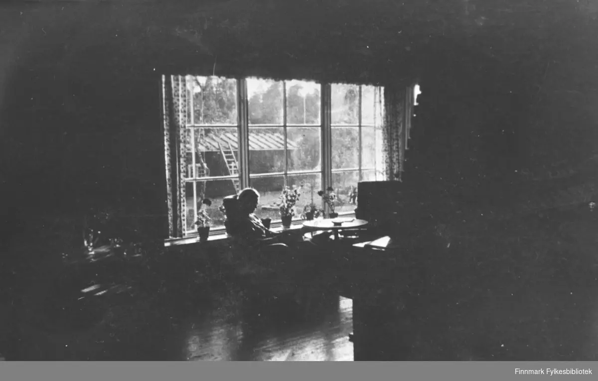 En kvinne sitter ved vinduet i et stort tømmerhus  i Petsamo / Petsjenga