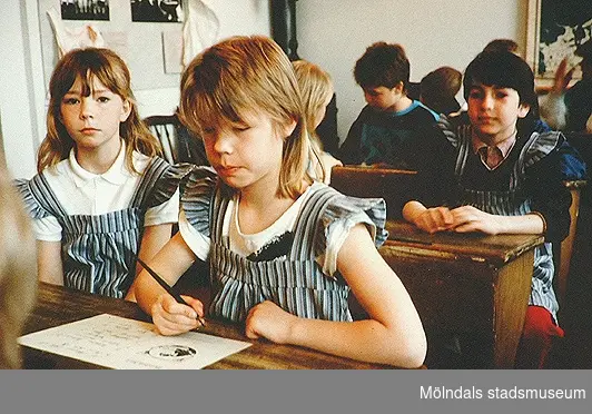 Barn som sitter i skolbänkar av äldre modell. Mölndals museum, 1990-tal.