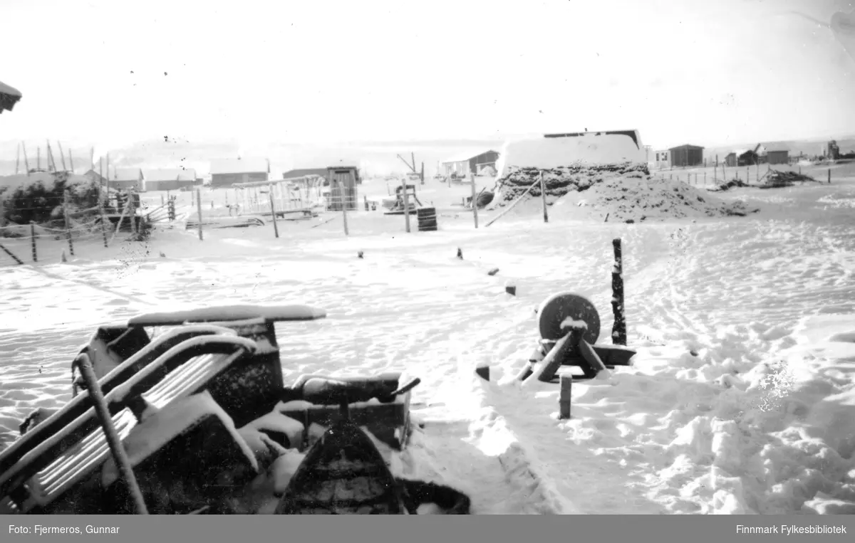 Bilde fra Karasjok en kald vinterdag i 1947. Noen sleder og en slipstein står foran i bildet og noen bygninger på andre siden av jordet.