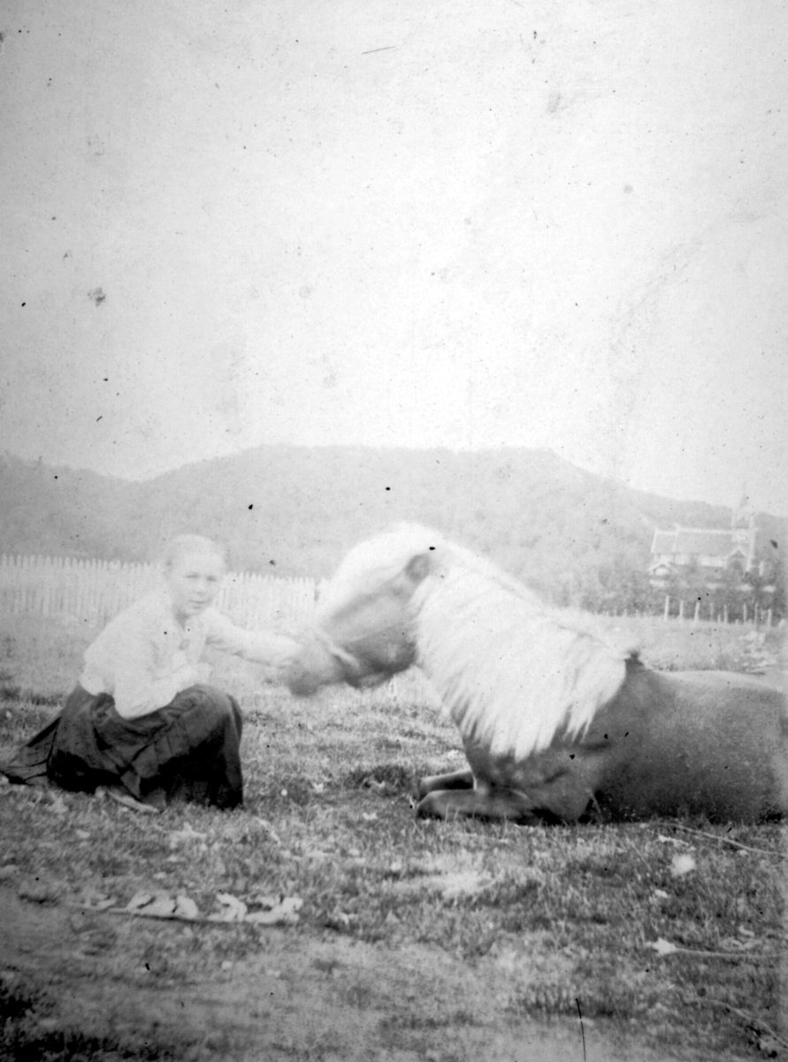 Andrea Håheim og hesten "Ola". Andrea klapper hesten som ligger på bakken. Til høyre i bakgrunnen kan man se en kirke.