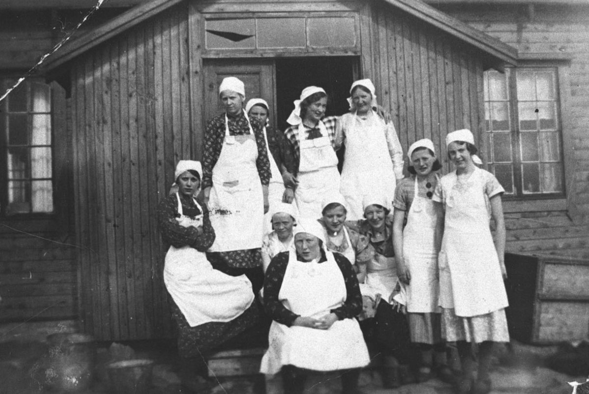 Jentene på husstellkurs fotografert på trappa ved et hus. Alle åtte unge kvinner og tre kvinner i 30-40 årene er kledd i hvite forklær og hvite tørkler.