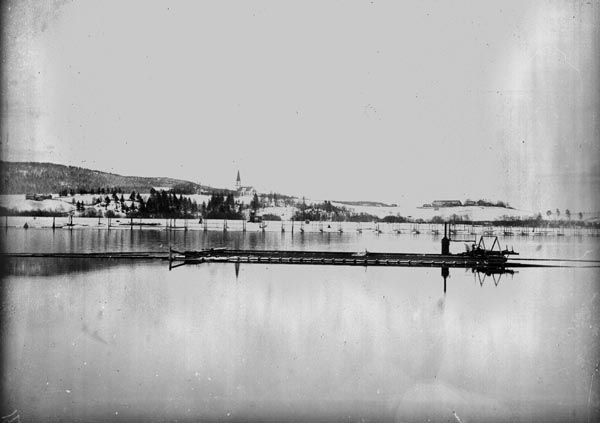 Den første soppemaskinen (moseapparatet) ved Fetsund lenser, fotografert nedenfor Leirtangsjunken i 1904.   Maskinen ble tatt i bruk påfølgende vår og var i drift i ti sesonger.  Soppemaskinene ble brukt til å bunte tømmer som skulle bukseres, i dette tilfellet over innsjøen Øyeren, med slepebåt som trekkraft.  Tidligere ble det brukt ringbommer når tømmeret skulle passere denne 34 kilometer lange innsjøen på veg mot nedre Glomma og industrien i Sarpsborg- og Fredrikstad-regionen.  Det var fløtingsfunksjonærer i Nedre Glomma som førte an i utviklinga av mosemaskiner for norske fløtingsvassdrag.  Pioneren var Richard Kristensen Furuholmen, som i 1876 fikk bygd en maskin ved Furuholmen lense som la tømmeret i tre lag - «flak» - med kryssende retning.  I 1901 lanserte kollegaen Arnt Svartedal en maskin som besto av to parallelle pontonger med ei faststående bru i den ene enden og ei skinnegående, bevegelig bru som kunne kjøres fram og tilbake.  Begge bruene hadde nedstikkende stålbein, som presset paralleltliggende tømmerstokker sammen, slik at det kunne slås vaiere rundt dem.  Soppemaskinen på fotografiet ser ut til å være bygd etter Svartedals prinsipp.  Til høyre, ved den faststående brua, ser vi noe som kan ha vært en liten dampmaskin som gav kraft til manøvreringa av den frittgående brua.  På bakkekammen i bakgrunnen ser vi Fet kirke og gardstunet på eiendommen Jaer.