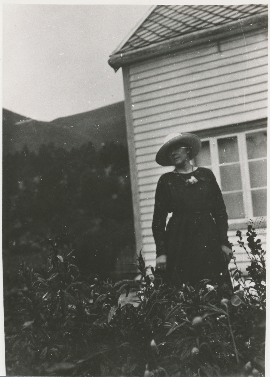 Dame med hatt i hagen foran et hus.