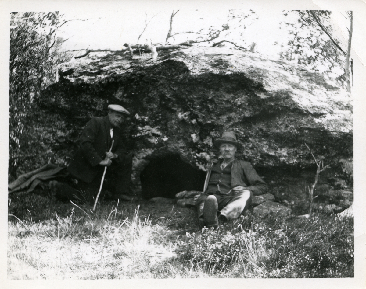 To menn sitter under en stein.
Steinen er sannsynligvis "Verssteinen" i Vang kommune.
