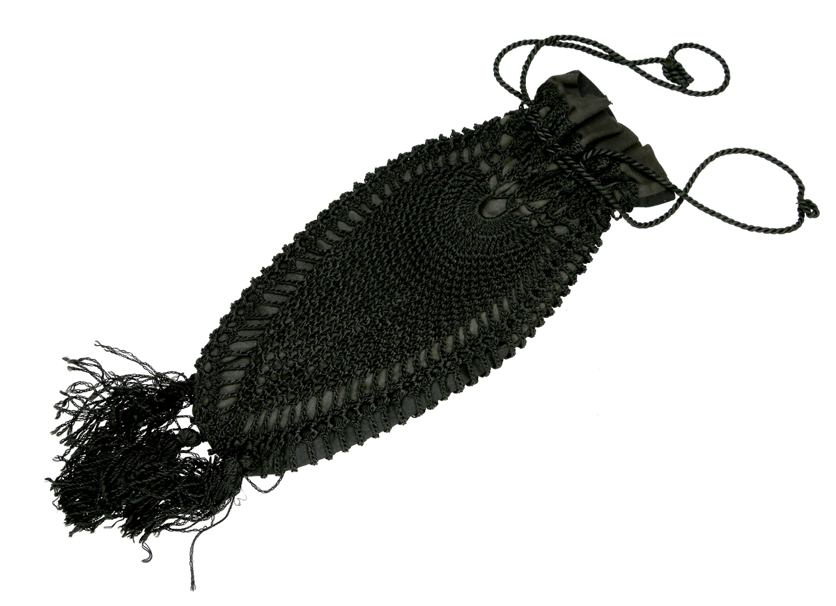 Veske utformet som en pose med knyting øverst, og heklet av svart tykk tråd med blank overflate. Vesken har et kypertvevd svart stoff på innsida av heklingen. Nederst er det dusker.