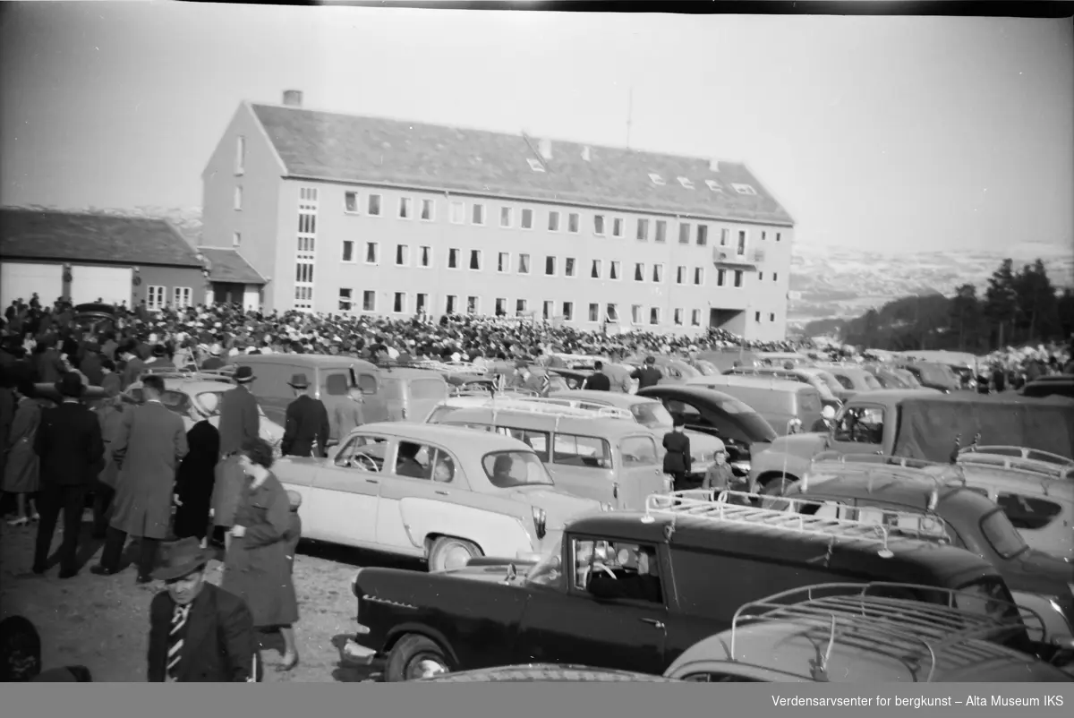 En stor folkemengde samlet utenfor et bygg på 17. mai i Alta. Biler står parkert nærmest kameraet.
Bildet er tatt på våren 1962.