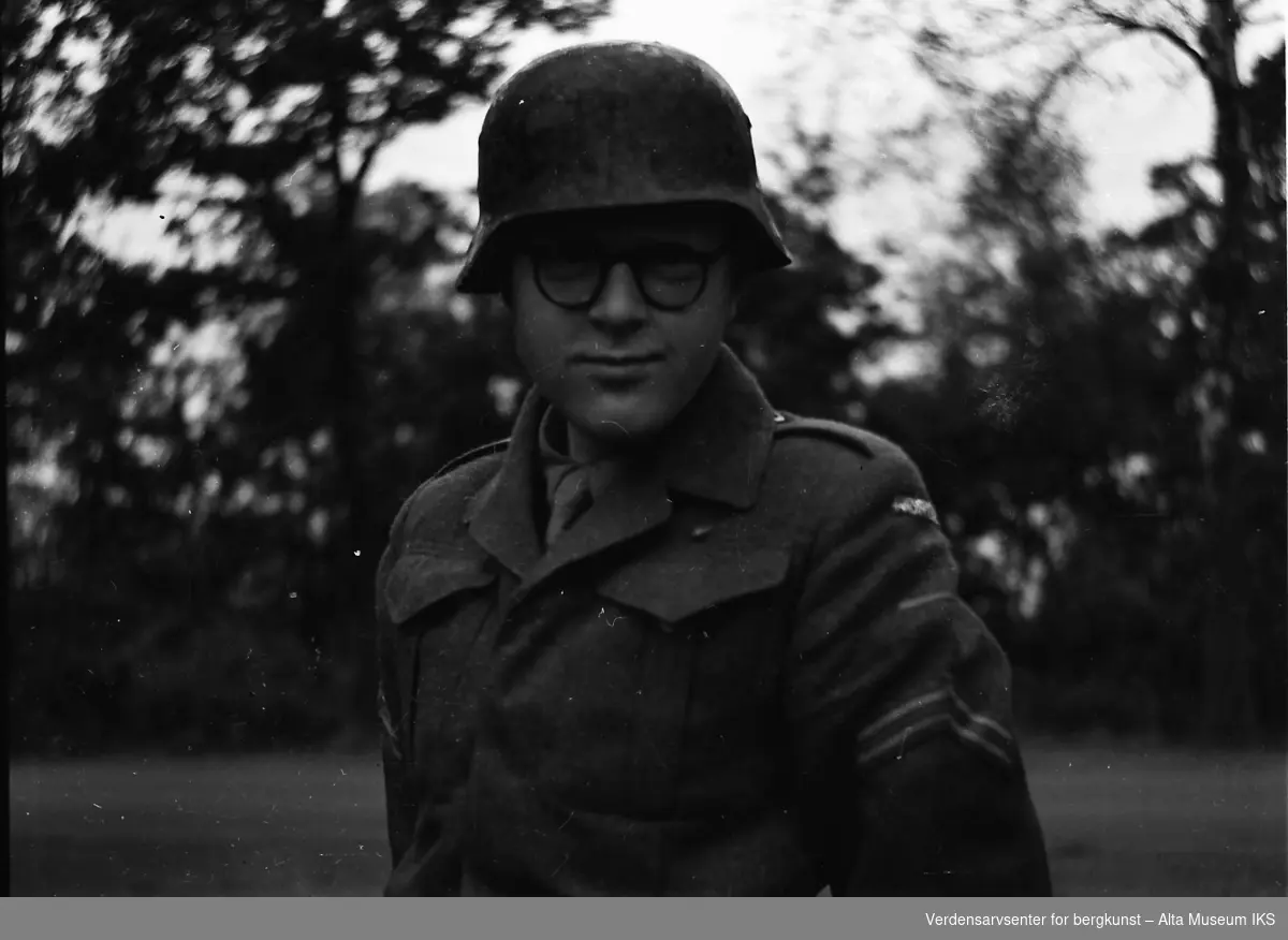 En ung soldat i uniform og hjelm står foran en skog. Han har et par runde briller på seg.