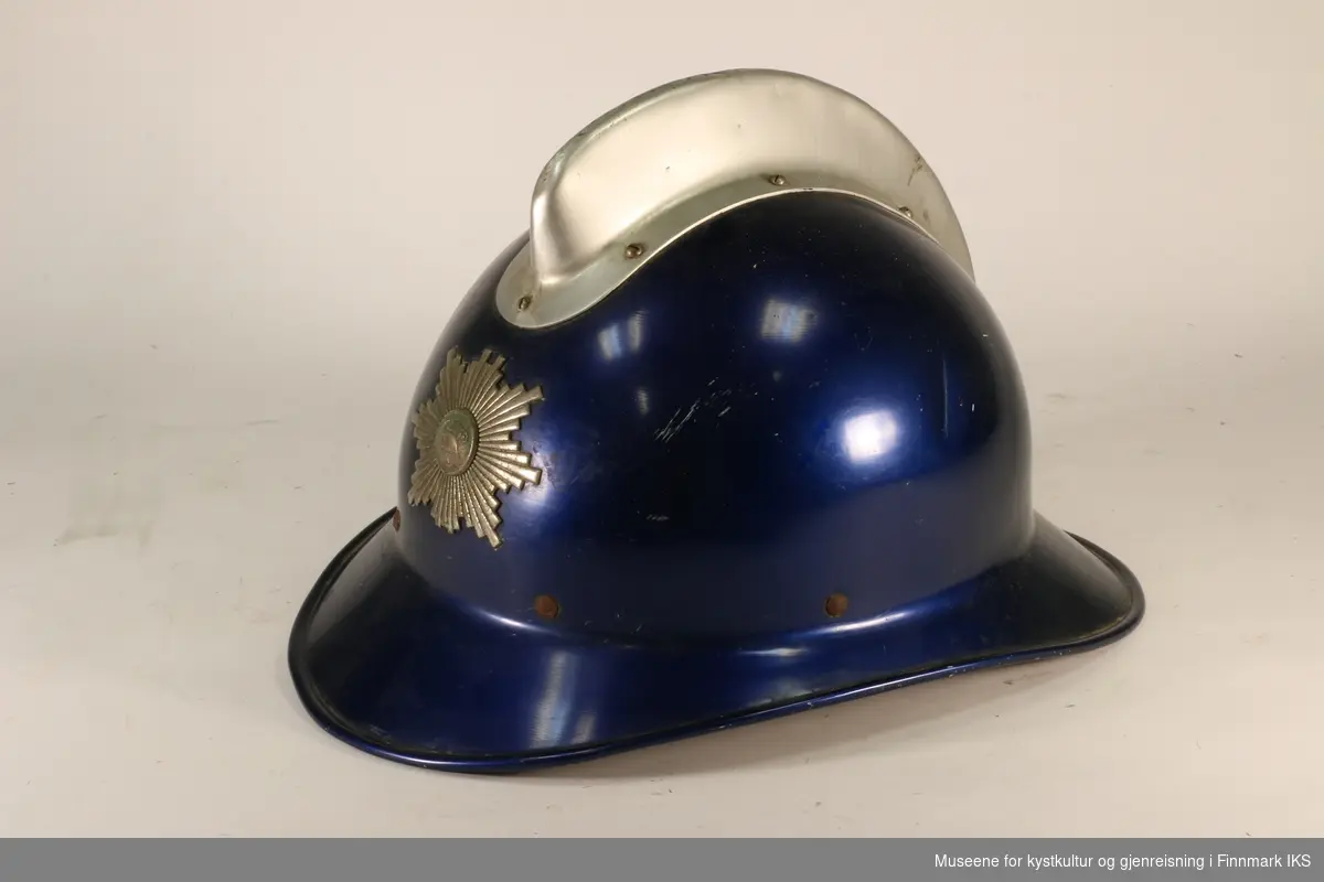 Blå brannhjelm av metall med støpt hanekam på toppen. Foran er det brannvesenets emblem. Innerfor av hjelmen er av skinn. Foret kan reguleres med snor.