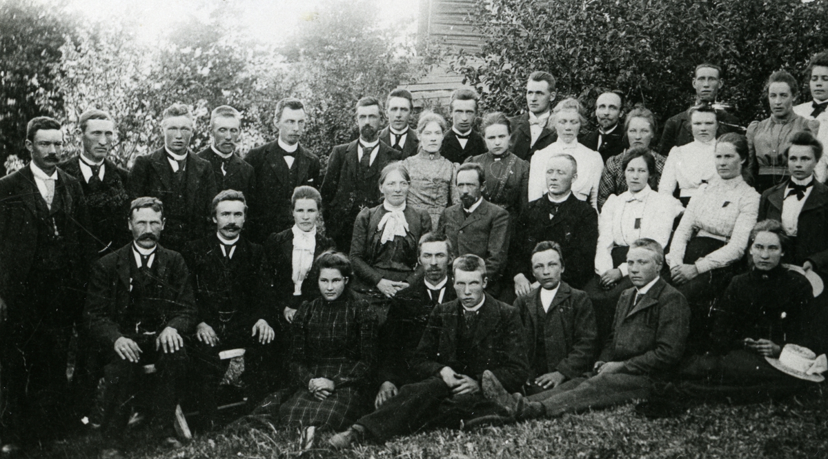 Gruppebilde av 33 kvinner og menn utnfor en bygning. Kvinnene er kledd i skjørt og bluser og karene har dress og slips eller sløyfe. Bildet er tatt ca 1900, Nordre Klevgård.
