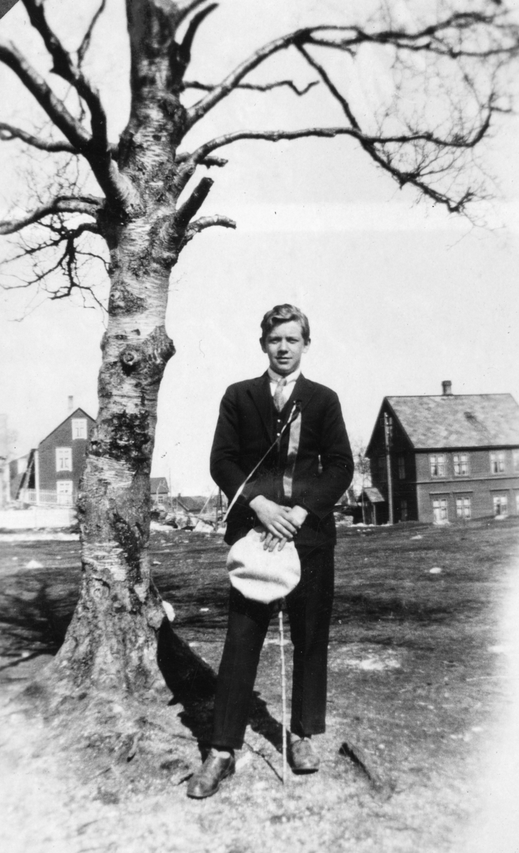 Ung mann ved et tre, bebyggelse i bakgrunnen.
