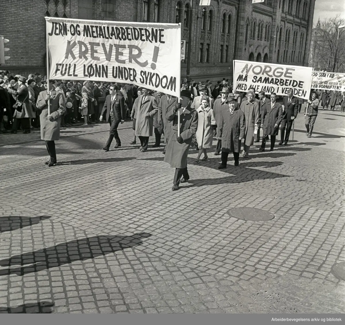 1. mai 1964 i Oslo.Demonstrasjonstoget i Karl Johans gate.Parole: Jern og metallarbeiderne krever!.Full lønn under sykdom.Parole: Norge må samarbeide med alle land i hele verden