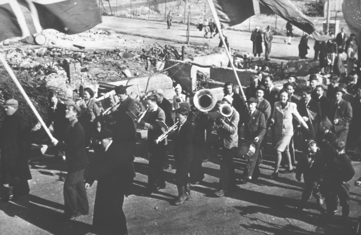 17. mai-tog i Kirkenes etter krigen. Et lite korps med trompeter, horn og tromme etterfulgt av smilende mennesker. I bakgrunnen ser vi ruiner etter bombinga.