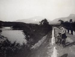 Kvinne står med sykkel på en vei. Vann til venstre.