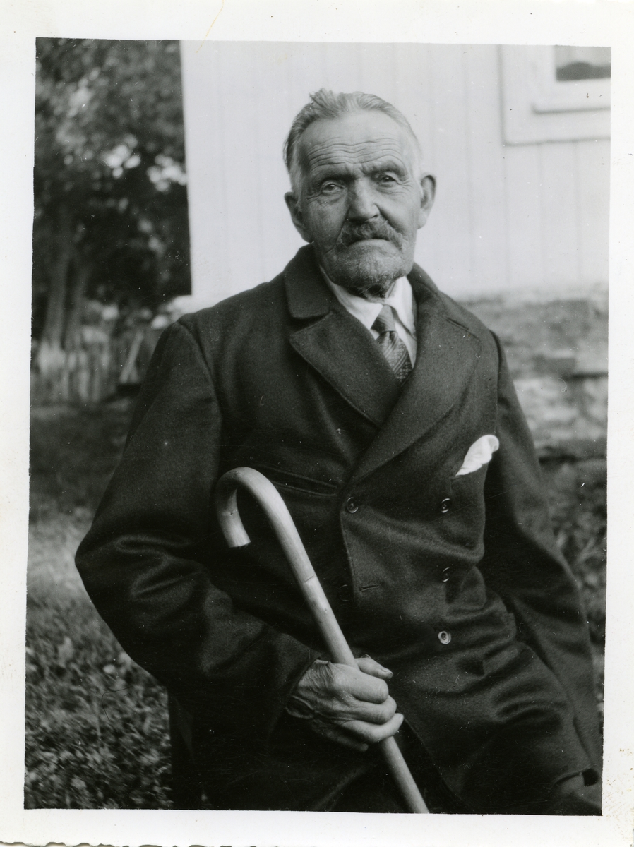 Portrett av en eldre mann sittende med en spaserstokk i hånda. Mannen er iført dress og skjorte med slips.