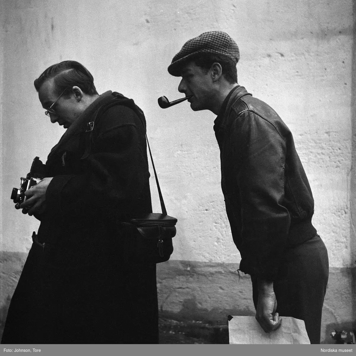Paris. Fotografen Rune Hassner tittar på sin kamera, iakttagen av ytterligare en man i keps som röker pipa.