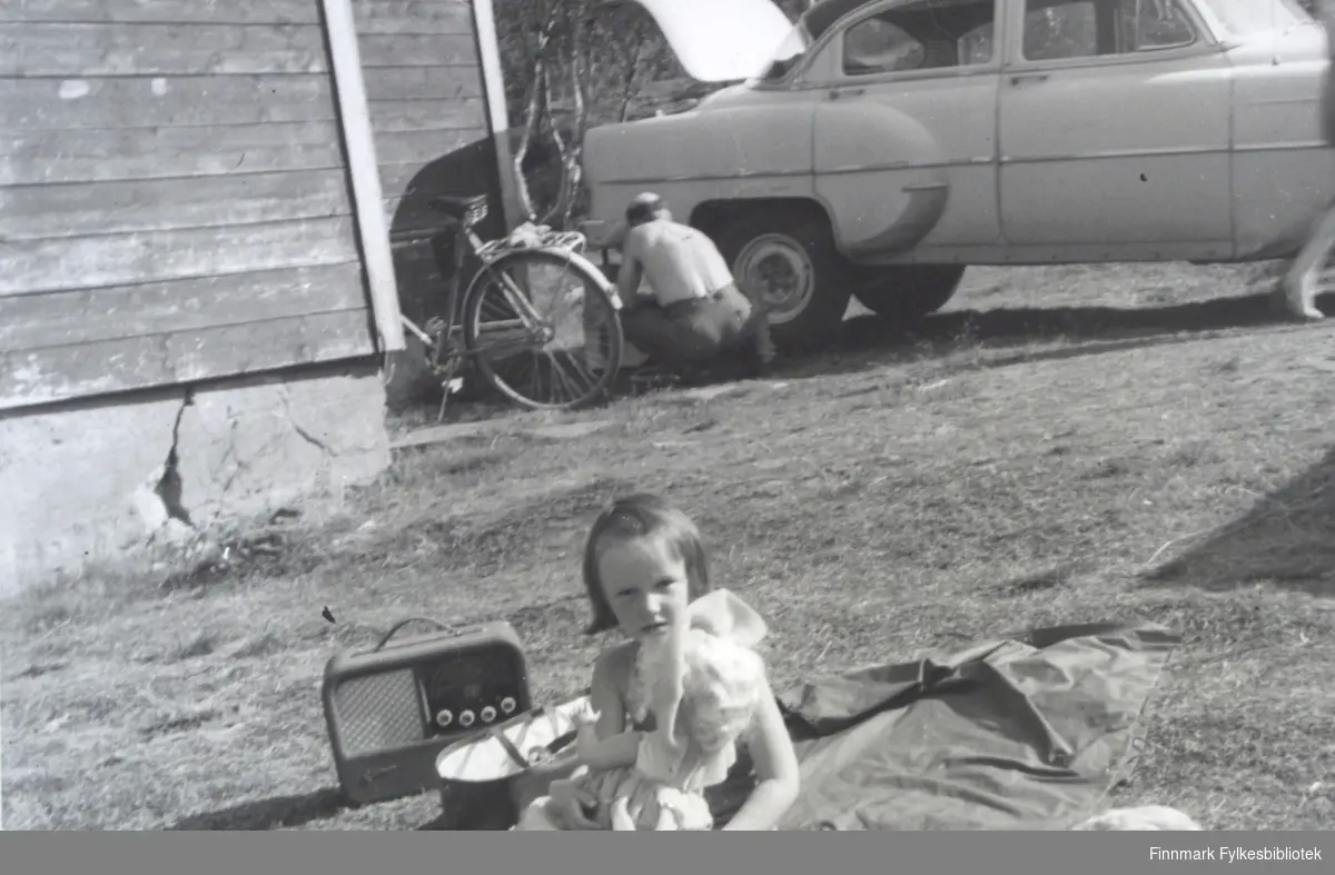 En sommerdag på Sælø familiens hytte ved Smalfjordvann i Tana ca. 1960. Slektninger er på besøk. Ei lite jente Gro Haldorsen leker med dukke og lytter på radio. Håkon Harøy jobber med en bil (Chevrolet 1953-54-modell) i bakgrunnen. Han var opprinnelig fra Bjugn og var formann på Vadsø Sildoljefabrikk.