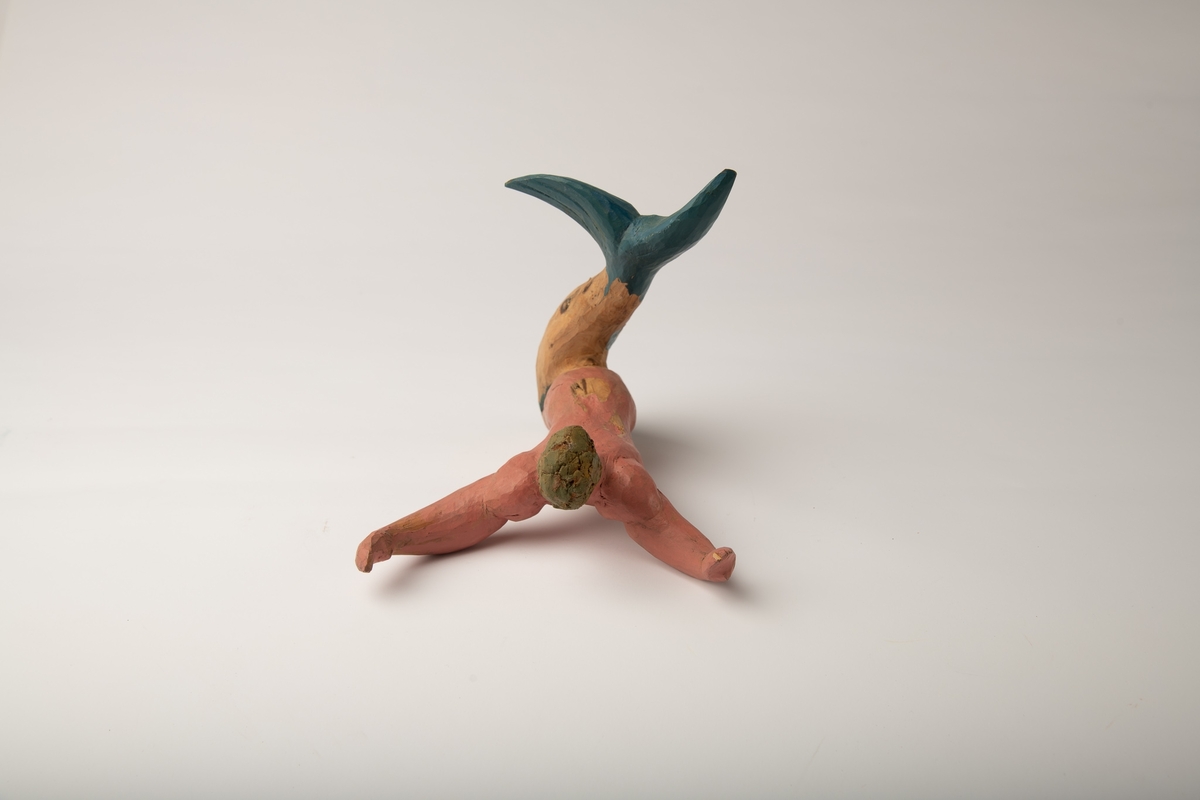 Mannsfigur i vinkel med blå halefinne og delvis rosa kropp. Hendene ut fra kroppen. Skåret av et stykke tre, der treets naturlige form har blitt en del av skulpturens form.