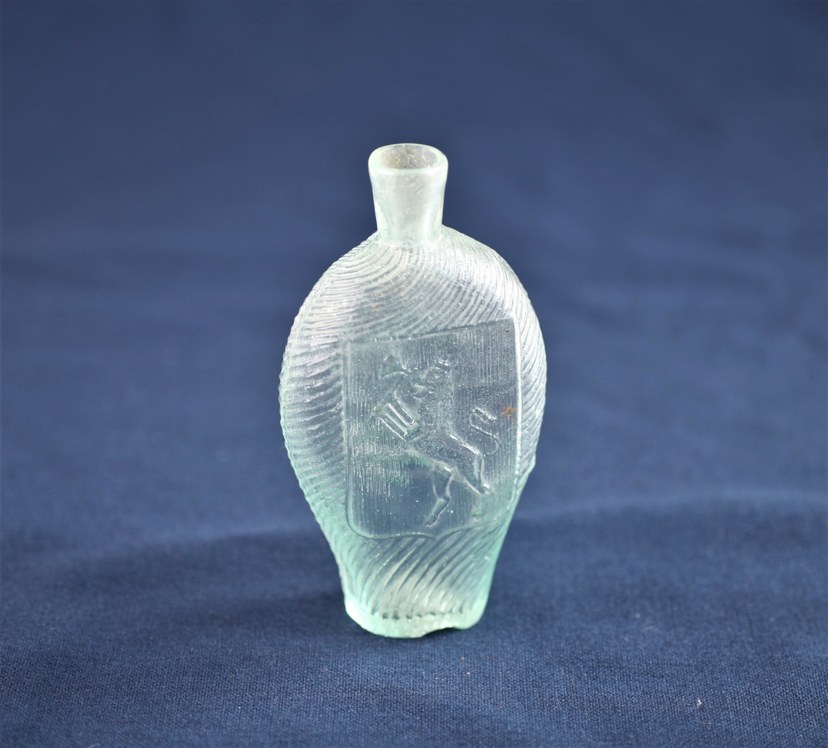 Flaska er av lyst grønt glass og har løvemotiv på den ene siden og en drueklase på den andre.
