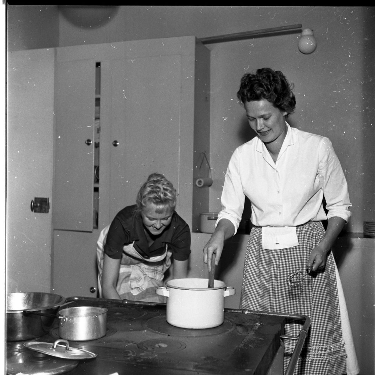 Två kvinnor vid en järnspis. Kvinnan till höger rör i en kastrull och kvinnan till vänster, som heter Birgit Roman, tittar i ugnen.
