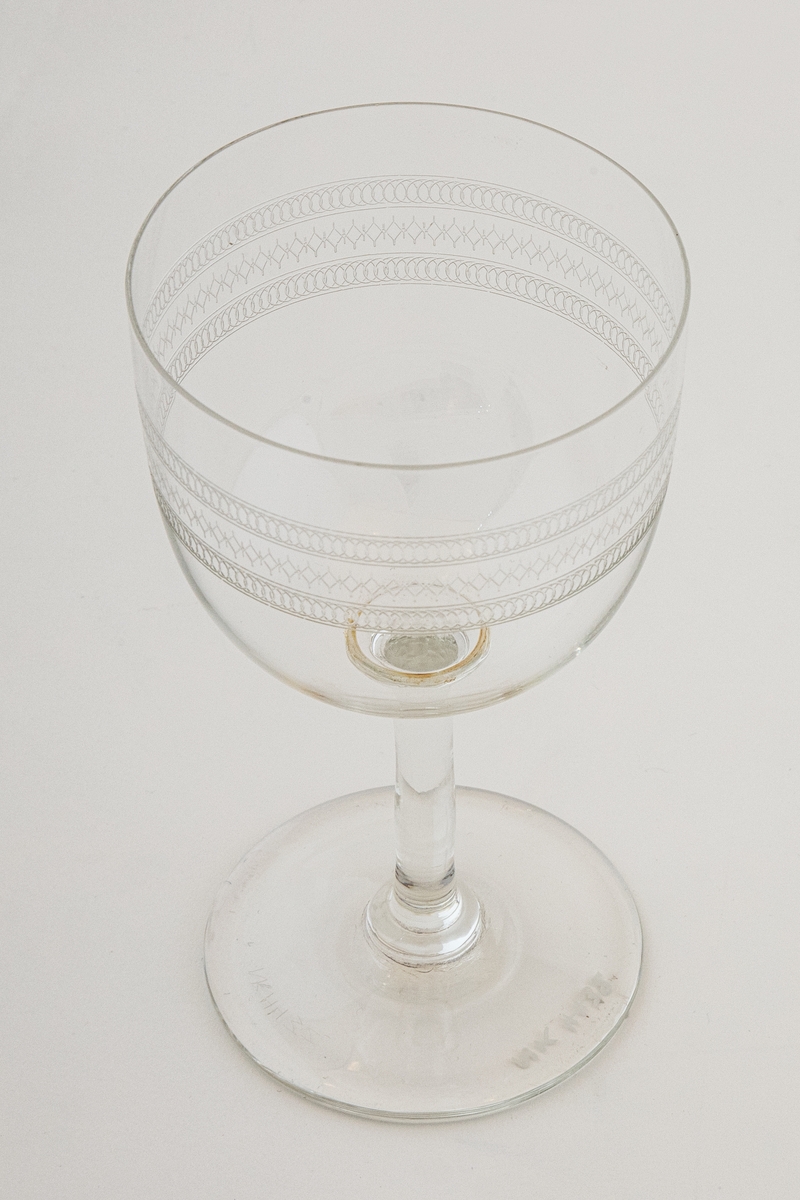 Et ølglass i klart glass, dekorert med etset og guillochert bord. Borden består av tre horisontale bånd med gjentagende geometriske figurer, i form av sirkel- og ruterformer. Klokkeformet kupa, hvor en ringformet vulst markerer overgangen til stetten. Sirkulær fotplate, noe hevet på midten.