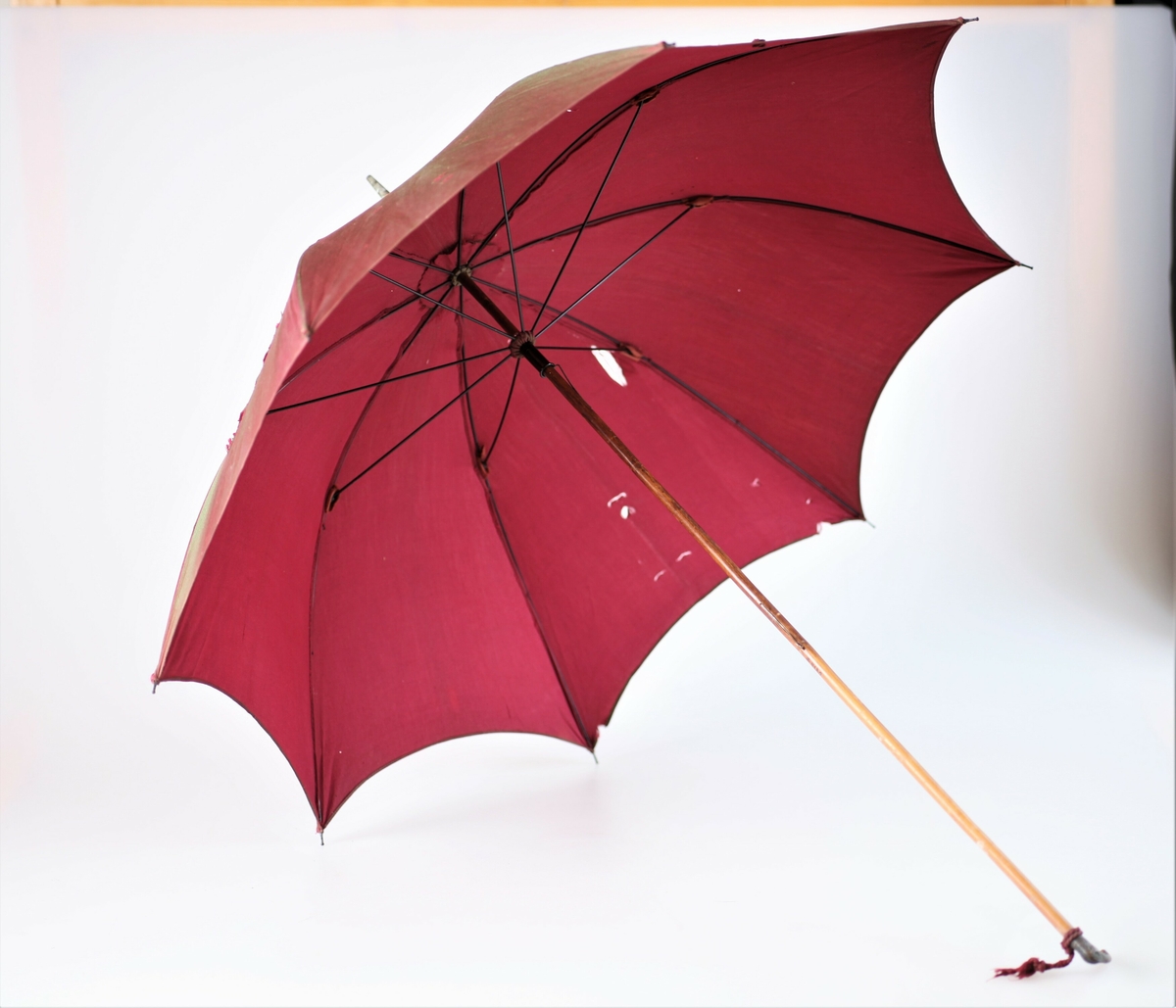 Paraplytrekket er rødlilla. På retten skinner stoffet i et grønnskjær. Dekorert håndtak av metall.