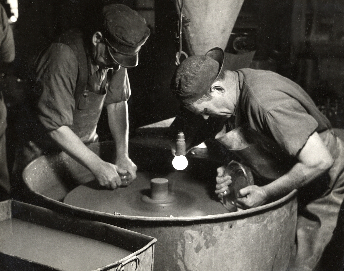 Orrefors glasbruk.
Två män slipar några glasföremål vid en slipsten. Till vänster Albin Bengtsson.