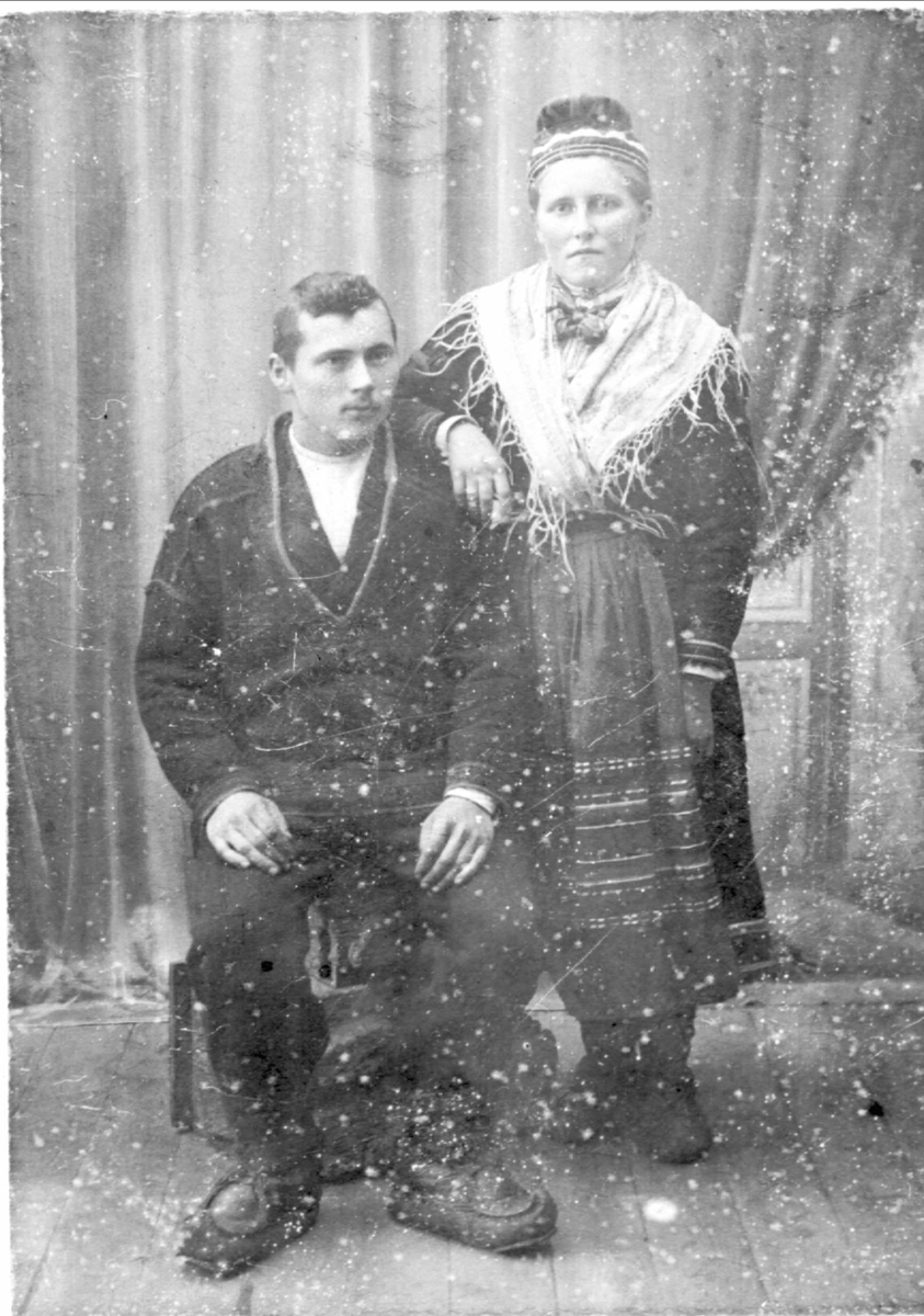 Studioportrett i helfigur av sittende mann og kvinne stående ved siden. Begge i samisk drakt.