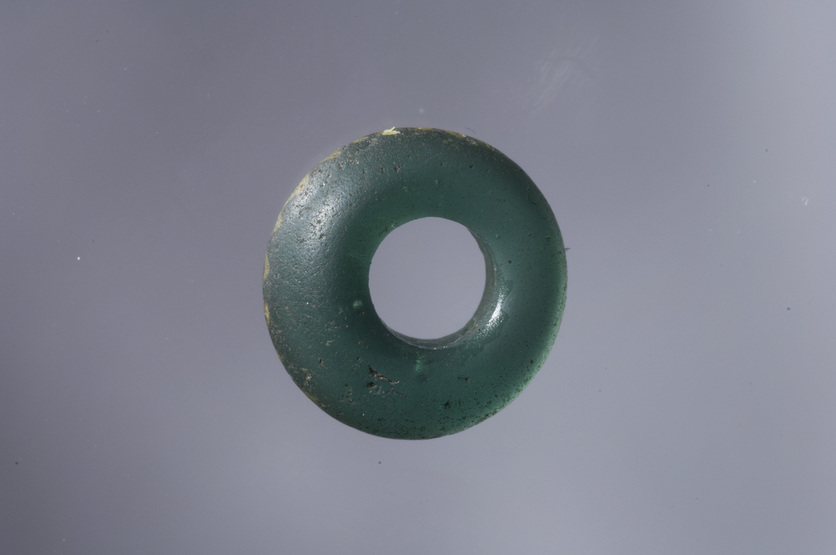 "Grønn ensfarget glassperle, 1,5 cm i tverrmål og 6 mm tykk." Ny beskrivelse etter revisjon: Perle med plane, ikke parallelle sider av grønt, gjennomskinnelig glass, dekorert med én rundtløpende gul sikk-sakklinje (Callmers perlegruppe Bc, tidligste type). L.: 5,5 mm - 7,8 mm, diam.: 15 mm. 