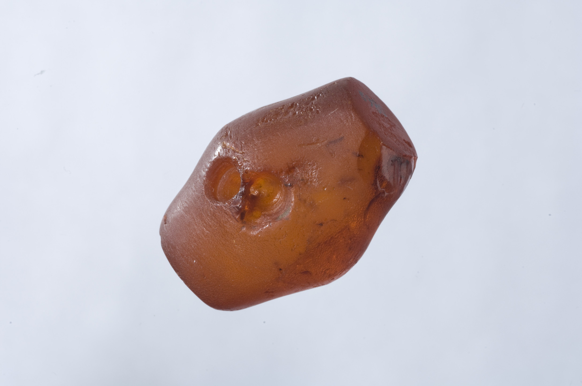 "Større perle av lys gulaktig rav fra stenalderen av dobbelt konisk form med rett avskårne ender. Midt på stykket er innboret et bærehull med V-formet boring. Perlens lengde er 3,3 cm., største bredde, på midten, 2,4 cm. - Sl. m. h. t. type Müller: Ordning Bronsealderen fig. 118." Ny beskrivelse etter revisjon: Perle av rødbrun rav, dobbelkonisk med rett avskårene ender. Bærehullet i samsvar med beskrivelsen. L: 33 mm, stdiam: 25 mm, endenes diam: 15 mm. Ett stykke av kanten er slått av i ene enden. 