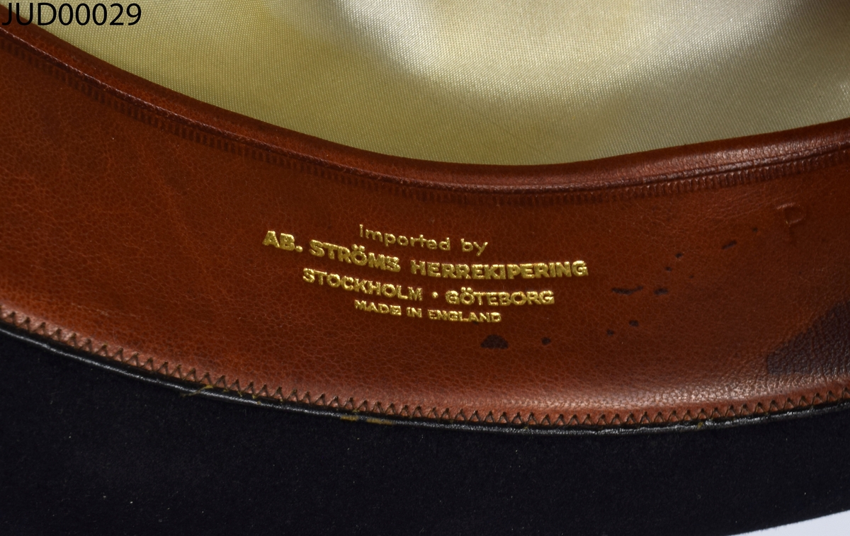 Hatt liggandes i sin originalkartong. Hatten är tillverkad av svart filt, med brunt läder och beige siden inuti.