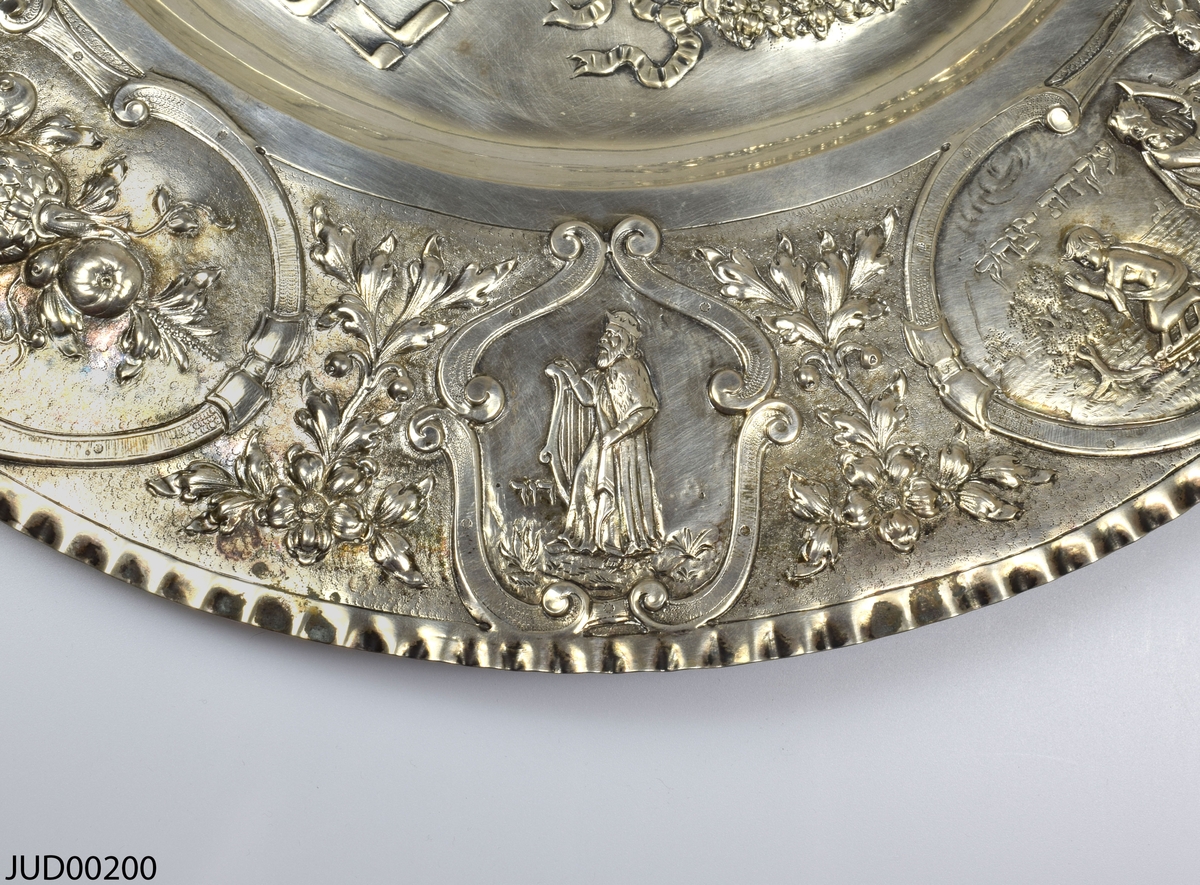 Stort silverfat tillverkat för pesach. Fatet är rikt dekorerat med präglad dekor i form av hebreisk text på spegeln samt blommor, frukt, personer (Aron, David, Mose) och olika scener på kanten. Vågig kant.
