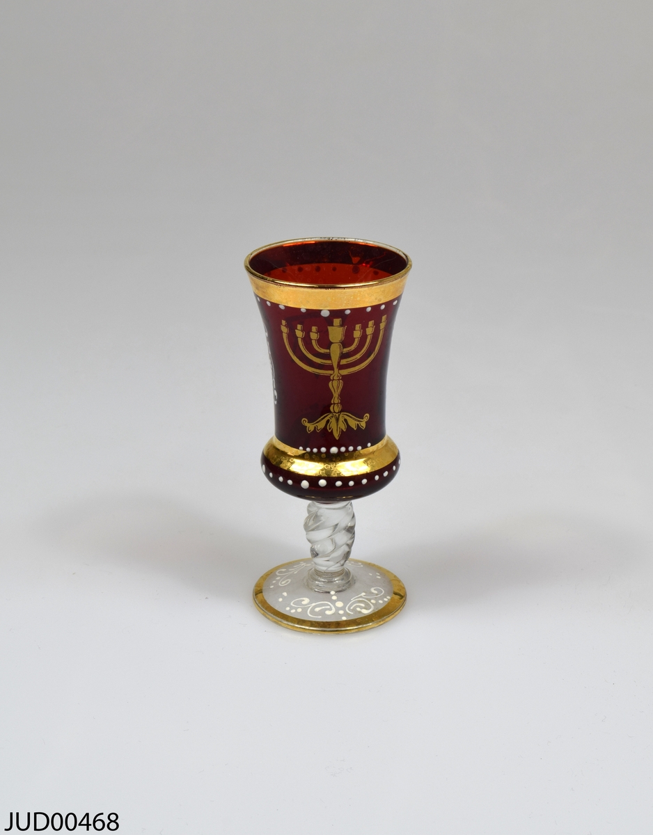 Glasbägare, med genomskinlig fot och rödfärgad cupa. Dekorerad med en guldfärgad menora och en davidsstjärna med hebreisk text i. Även vit snirklig dekor.