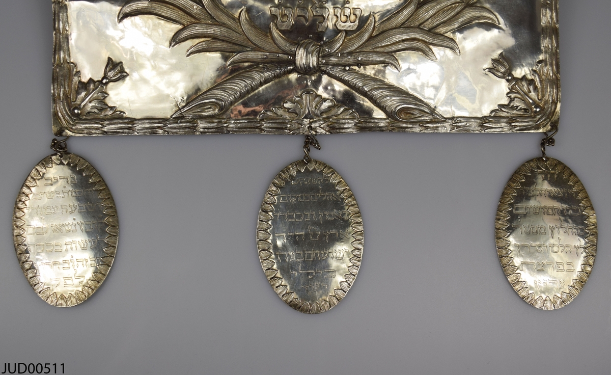 Torahsköld tillverkad av silver. Den här svenska stämplar i form av O2, kattfot, St Erik och EB. Dekorerad med lejon som flankerar en krona och sköld med hebreisk text. Tre hängande plaketter med ristad hebreisk text i nederkant.