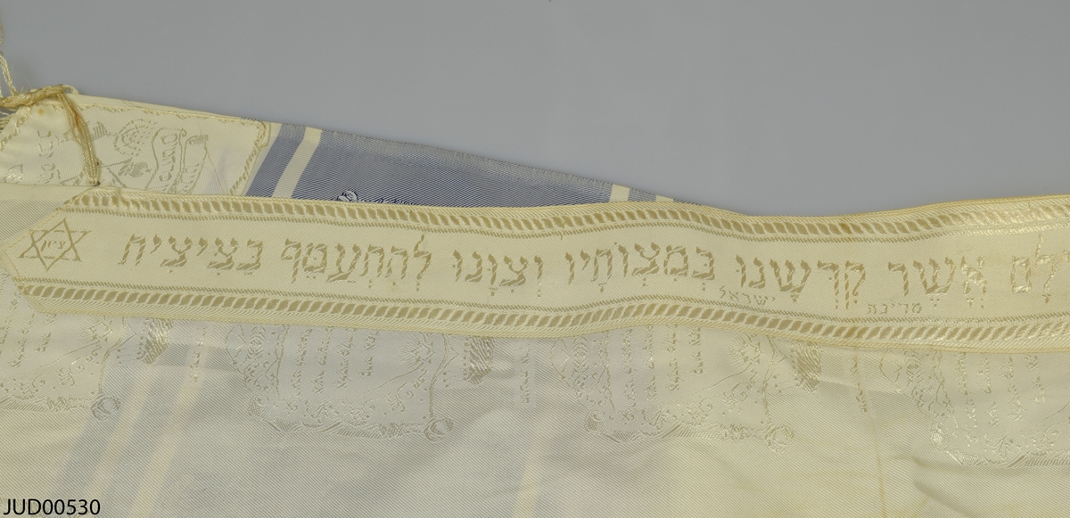 Bönesjal, troligtvis tillverkad av maskintillverkad satin. Invävd dekor i form av blå ränder med infällda sköldar i vilka hebreisk text är skriven. Likdana sköldar i vitt på det vita. I ena kanten finns ett band med hebreisk text.